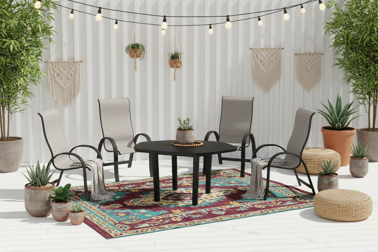 This is a lifestyle image showing this patio dining set on a white balcony. There are potted plants, jute ottomans, hanging lights and a colorful outdoor rug.