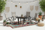 This is a lifestyle image showing this patio dining set on a white balcony. There are potted plants, jute ottomans, hanging lights and a colorful outdoor rug.