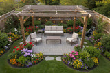 This is a lifestyle image taken from above. It's a beautiful backyard with lots of grass and flowers and a waterfall feature. The patio set is underneath a wooden pergola on a stone patio.