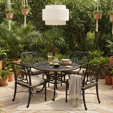 This is a lifestyle image showing this patio dining set on a stone patio surrounded by Midwestern foliage and potted plants. The frames of the furniture are black and the cushions are a light tan color. 