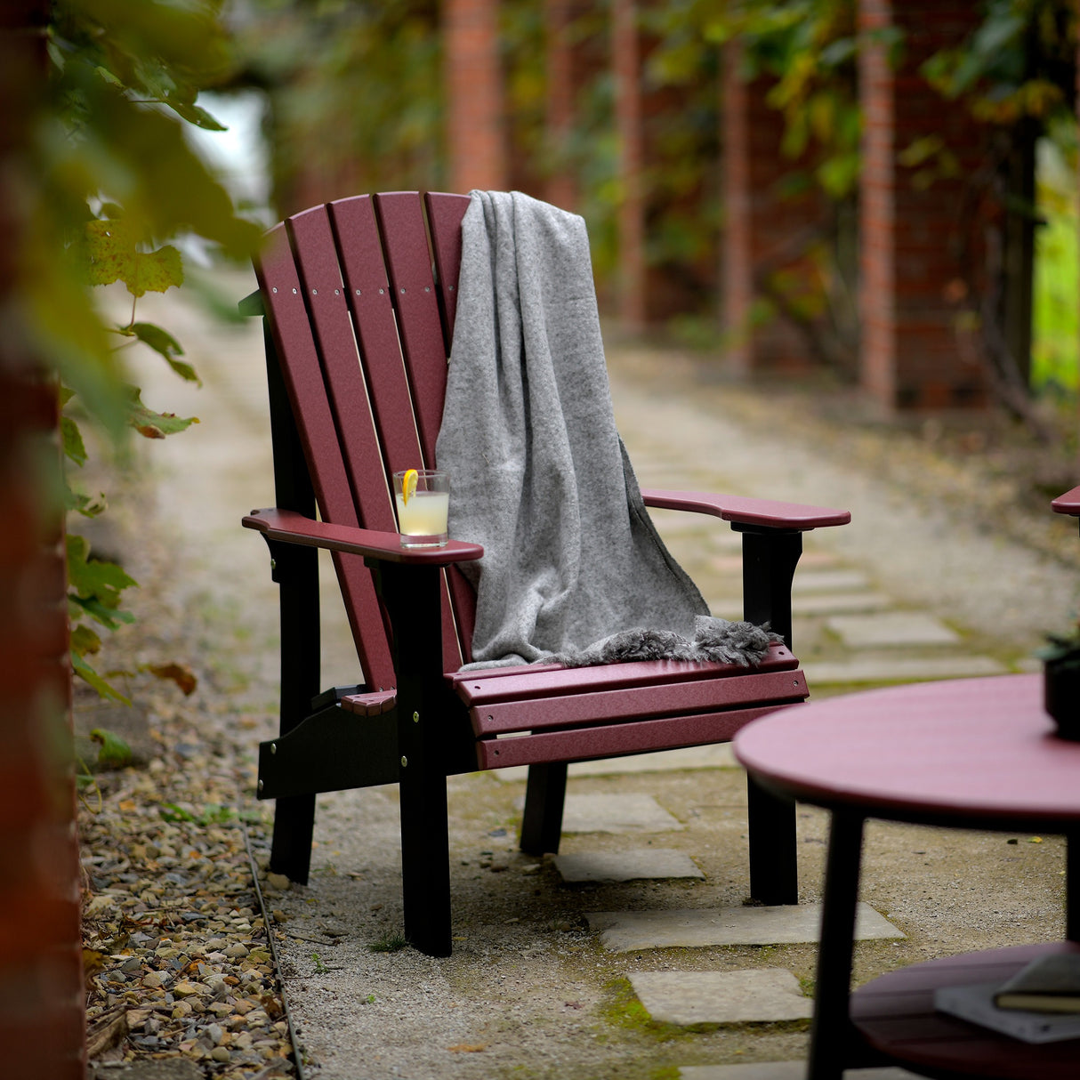 This is a lifestyle image showing the Cherrywood and Black colored Adirondack chair on a stone pathway in front of a matching table. There's a gray blanket draped over the side of the chair and a glass of lemonade on the armrest.