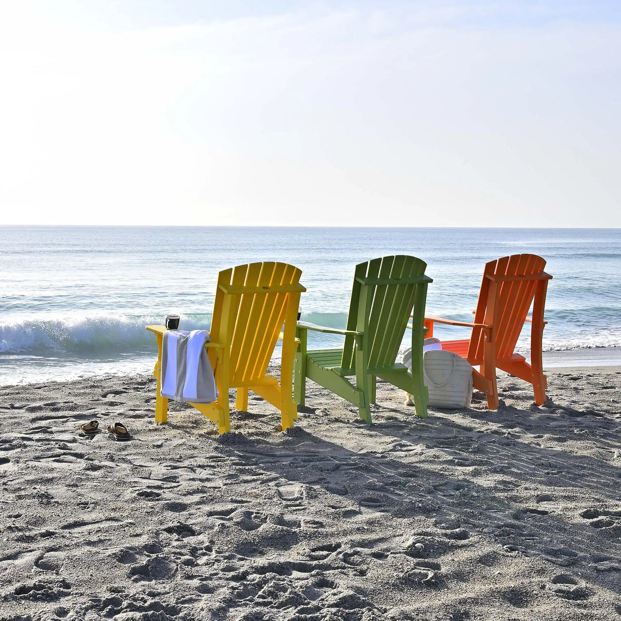 This is a lifestyle image showing three Adirondack chairs facing the ocean on a beach. One chair is Tangerine, one chair is Lime Green and one chair is Yellow.