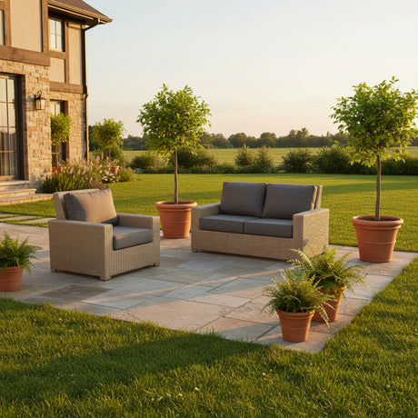 This is a lifestyle image showing the lounge chair and the loveseat on a stone patio in a grassy backyard. There's a house is off to the left. The furniture is surrounded by potted ferns and potted trees.