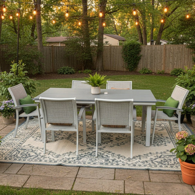 This is a lifestyle image showing this patio dining set on a stone patio in a grassy, Midwestern backyard. There are potted plants and hanging lights.