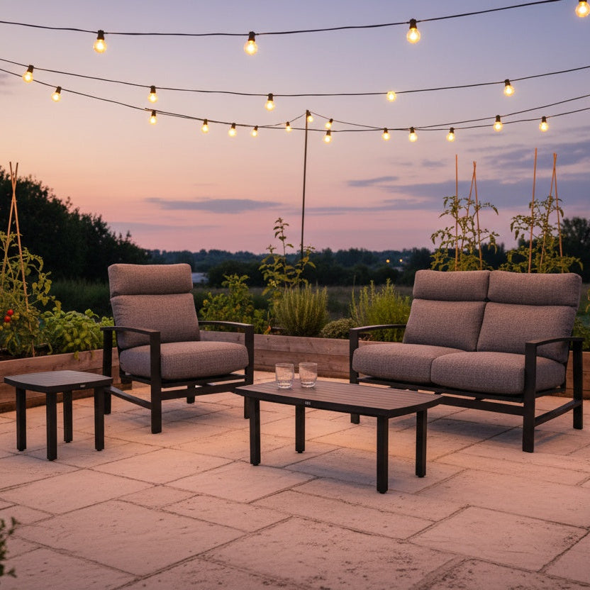 This is a lifestyle image showing this cushioned patio furniture set on a stone patio next to a garden at sunset. There are hanging lights above the patio.