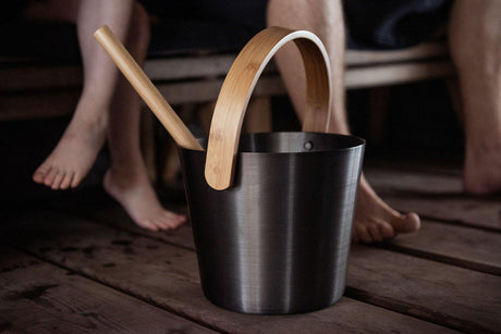 This lifestyle image shows the black aluminum sauna bucket on the floor inside of a sauna by some feet.
