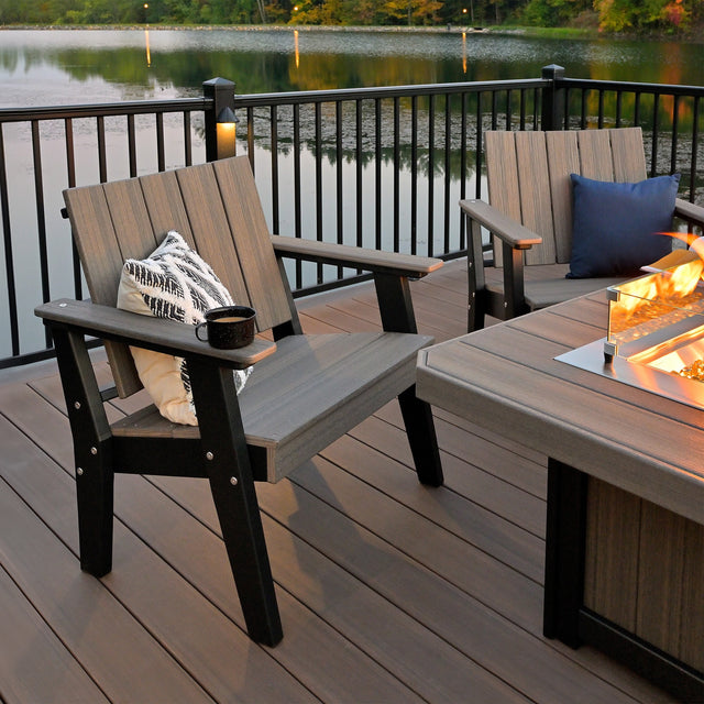 This is a lifestyle image showing two Coastal Gray and Black colored Urban Chat Chairs in front of a matching fire pit table on a balcony overlooking a small body of water.