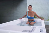 Long, rectangular tub designed for a cold plunge. The cabinet color is a tan-ish gray color and the inside is white. There's one seat. This image shows a woman sitting above the water, getting her legs acclimated to the cold.
