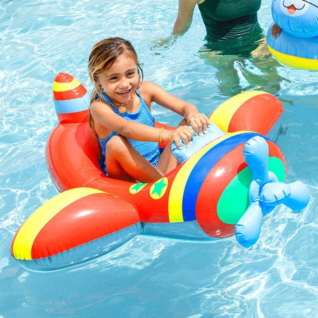 A little girl is sitting inside the airplane pool float in a pool. The float is red with yellow, blue and green stripes. The sides are decorated with yellow circles with green stars inside.