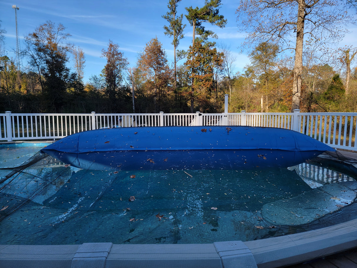 This is a lifestyle image showing the giant Pool Winterizing Pillow under a net cover on a large backyard pool. This image is taken from the side to show how wide the pillow is.