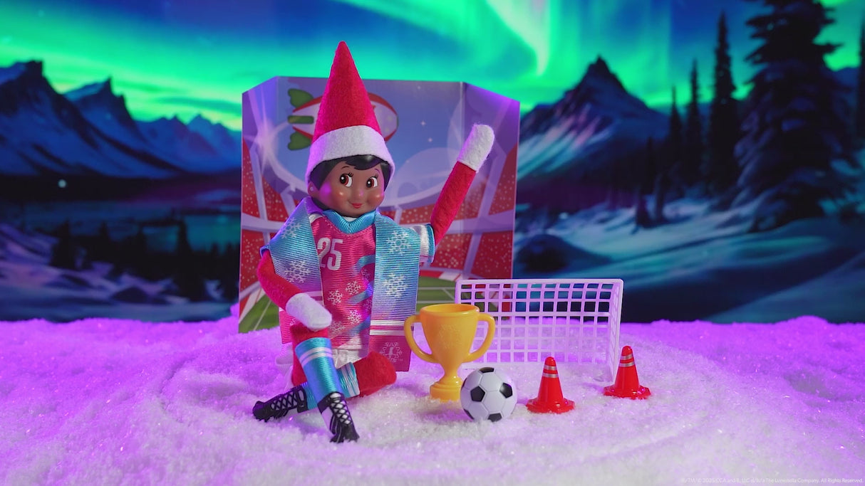 This video shows a Scout Elf sitting in front of the backdrop, next to a soccer net, the trophy, the soccer ball and two cones. He's wearing the uniform an the scarf.