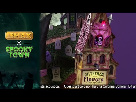 Resin building facade for a Spooky Town Lemax display. Designed to look like a bunch of store fronts. One section is a mortuary with a "help wanted" sign hanging from it. The building next to it reads "NOTION'S POTIONS" and has a witch hat for a roof. The building next to it is a flower shop. Behind them all is a cemetery and a spooky cathedral. There's a witch flying near the moon with bats following behind.