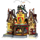 Porcelain lighted building for a Lemax display. Designed to look like a rustic cabin covered in snow and decorated for Christmas. There are angels and pigeons flying above. Santa is waving from a window in the attic. There are reindeer going in a circle around a decorated Christmas tree. There are ornaments for sale on the sidewalk and a nutcracker standing guard at the front door.