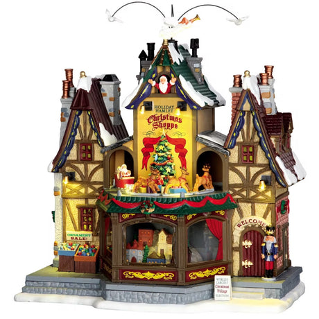 Porcelain lighted building for a Lemax display. Designed to look like a rustic cabin covered in snow and decorated for Christmas. There are angels and pigeons flying above. Santa is waving from a window in the attic. There are reindeer going in a circle around a decorated Christmas tree. There are ornaments for sale on the sidewalk and a nutcracker standing guard at the front door.