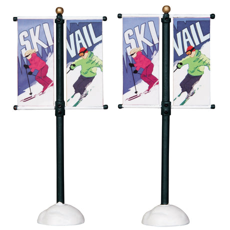 Plastic village accessories for a Lemax display. Designed to look like two street poles with banners on them. One side of the banner says "SKI" and features a woman skiing wearing pink winter clothes. The other flag reads "VAIL" and features a person skiing wearing a lime green hoodie, purple pants and a red hat.