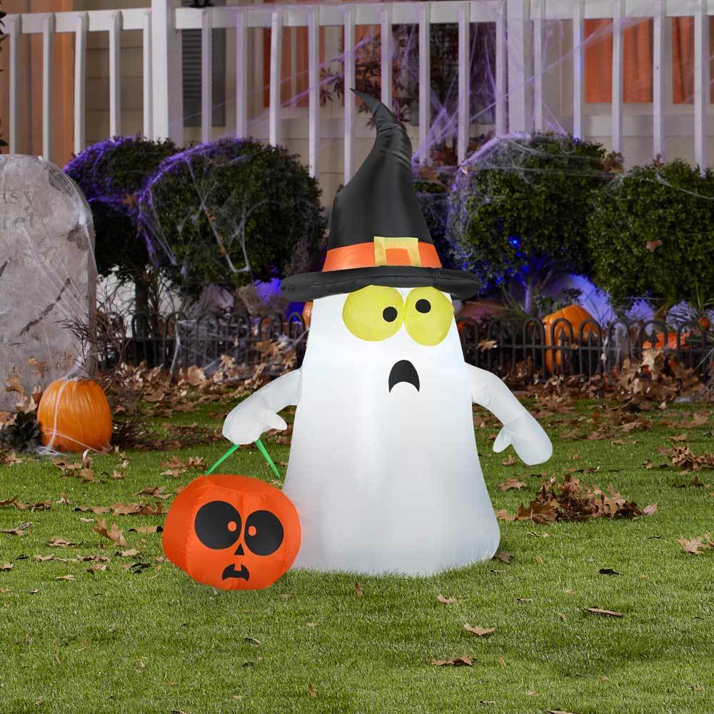 This is a lifestyle image showing the ghost inflatable in front of a house decorated for Halloween. The ghost is wearing a black witch hat with orange accents. He has yellow eyes and is holding a jack-o-lantern.