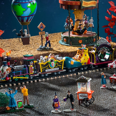 Polyresin train on a set of tracks for a Lemax display. The train cars are all different. The head of the train is the face of a clown. The second one is full of silver and wooden barrels and the side reads "Stitches the Clown: Traveling Mayhem." The third car is shaped like a giant clown shoe. The last one is shaped like a carnival ride where it spins you in a circle.