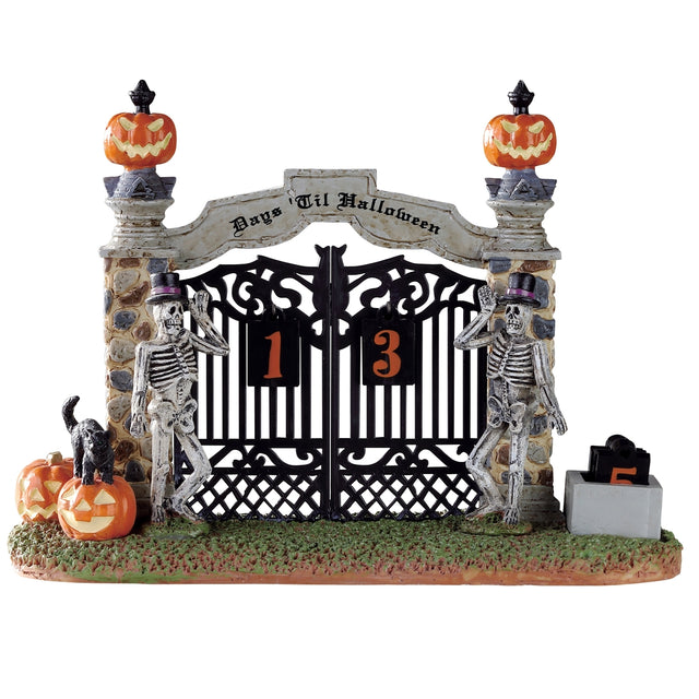 Polyresin village accent for a Lemax display. Designed to look like a metal gate with two stone columns. There are two skeletons wearing top hats standing in front of the columns. There are carved pumpkins and a black cat. It's a Halloween countdown.