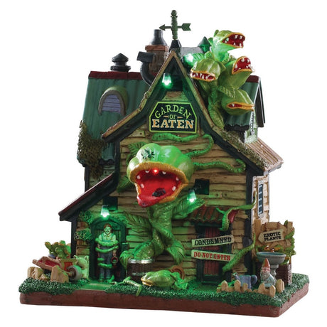Porcelain lighted building for a Lemax display. Designed to look like a cabin overrun with Venus fly traps with teeth. There are roots holding a person hostage at the front door. There's an elf hiding behind a little fence in the front yard. The lights are glowing an eerie green color.