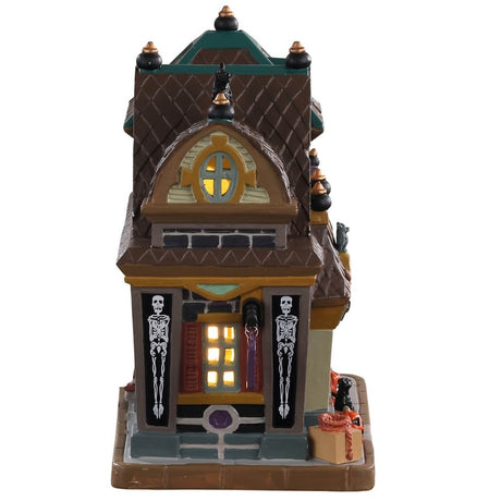 Porcelain lighted building for a Lemax display. Designed to look like a large shop. There's a black cat on the roof and carved pumpkins near the front door. There are skeleton accents and a bat near the front door.