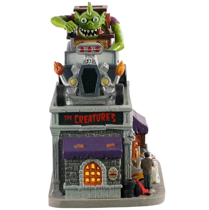 Polyresin and porcelain building for a Lemax Spooky Town display. Designed to look like a mechanic who does custom paint jobs for cars. There's a large green monster on the top, driving a giant car shaped like a house. The sign on the sign of the building reads "THE CREATURE'S."