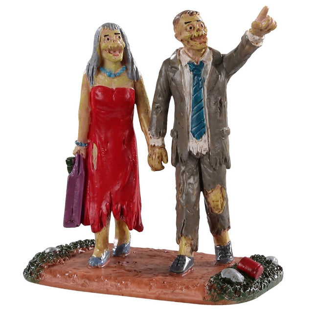 Polyresin figurine for a Lemax Spooky Town display. Designed to look like two spooky monster people wearing raggedy clothes. The woman monster has long gray hair and is wearing a red ripped up dress with blue jewelry and shoes. She's holding a purple bag. The man monster is wearing a torn up gray suit with a blue tie. They're walking on a dirt path with grass on the sides. The man is pointing at something far away.
