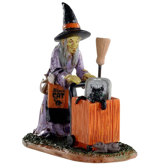 Polyresin figurine for a Lemax Spooky Town tabletop display. Designed to look like a smiling, old witch pushing a cart with a tombstone, a broom and a black cat inside. There's a rat in front of the cart. The witch is wearing a light purple dress, a traditional black and orange witch hat and an orange purse.