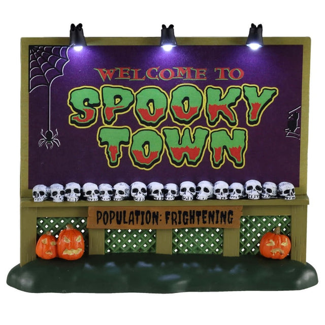Plastic village accessory for a Lemax Spooky Town display. Designed to look like a town sign that reads "WELCOME TO SPOOKY TOWN." There are skulls lined up at the bottom of the sign. The sign also reads "POPULATION: FRIGHTENING." There are three jack-o-lanterns on the grass under the sign. The sign also has three lights on the top. The sign is purple, lime green, red, black, olive green and brown.