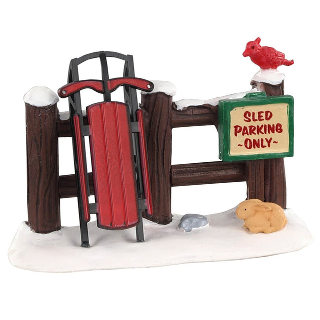 Polyresin figurine for a Lemax Village tabletop display. Designed to look like a sled up against a wooden fence with a sign on it that reads "Sled Parking Only." The fence and the ground are covered in snow. There's a red bird on the fence and a bunny on the ground