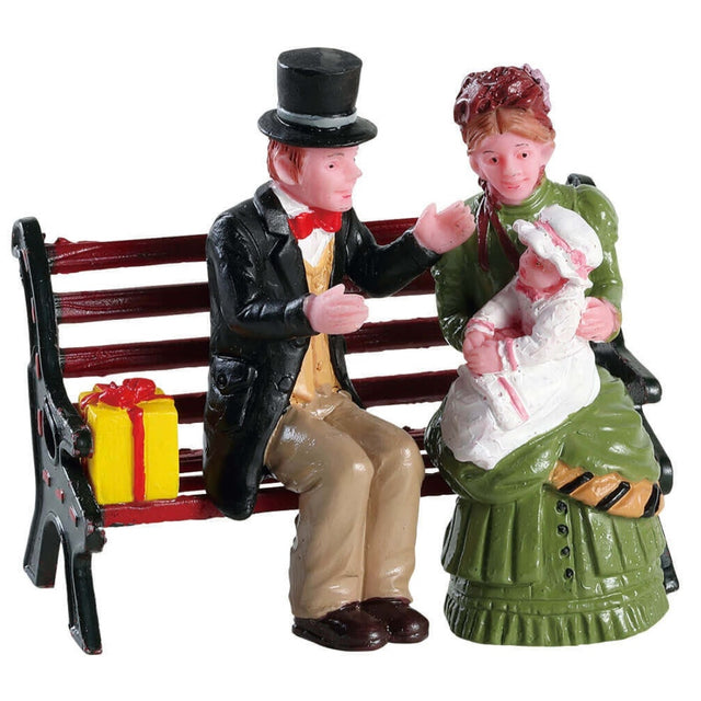 Lemax village accessory designed to look like a man and a woman in Victorian era clothes, holding a baby and sitting on a bench. There's a yellow and red present on the bench next to them.
