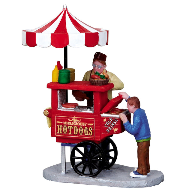 Polyresin figurine for a Lemax display. Designed to look like a hot dog vendor. A young boy is looking into a cooler that reads "Cold Drinks." The vendor is building the hot dog for the child. The cart reads "Delicious Hot Dogs" and is a bright red color. There are condiments on top of the cart along with a red and white striped umbrella.