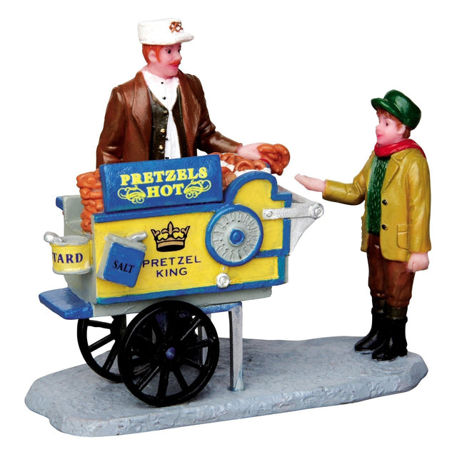 Polyresin figurine for a Lemax Village display. Designed to look like a pretzel vendor handing a pretzel to a young boy. They're both wearing winter clothes. The pretzel cart reads "Pretzel King" and features mustard and salt.