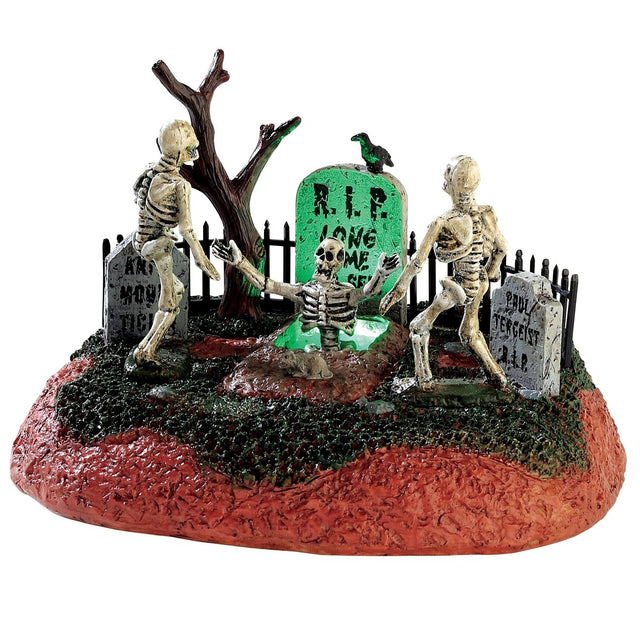 Polyresin table accent for a Lemax display. Designed to look like a graveyard with tombstones and a bare tree. There are two skeletons trying to help another skeleton out of a grave. There's a glowing green light coming out of the grave.