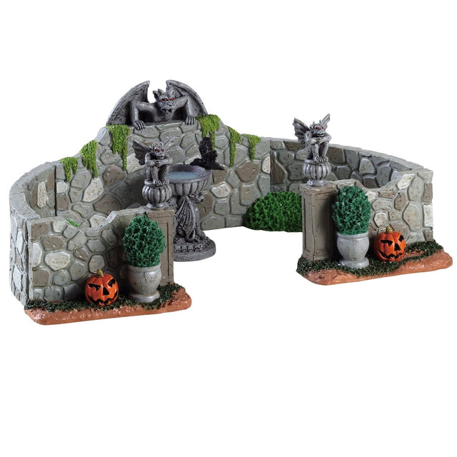 Polyresin village accent for a Lemax display. Designed to look like a stone fence with gargoyle accents. There's a birdbath with a crow in it and potted plants. There are two jack-o-lanterns on the ground near the entrance.