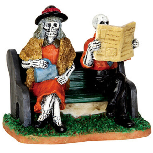 Polyresin figurine for a Lemax display. Designed to look like two skeletons sitting on a bench. The woman is wearing a fur coat and a red dress. The man is wearing a red shirt and black pants and is reading a newspaper.