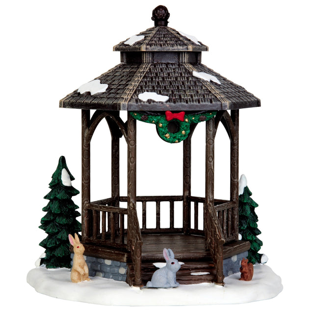 Polyresin table accent for a Lemax display. Designed to look like a wooden gazebo. It's got snow on the roof and on the ground. There's a decorated wreath with a red bow on top under the roof of the gazebo. There are two snow covered trees and features two bunnies and a squirrel.