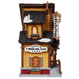 Porcelain lighted building for a Lemax display. Designed to look like an old Victorian-era house with wreaths and snow on it. The house is gold with brown frames. Inside the window you can see an array of chocolates.