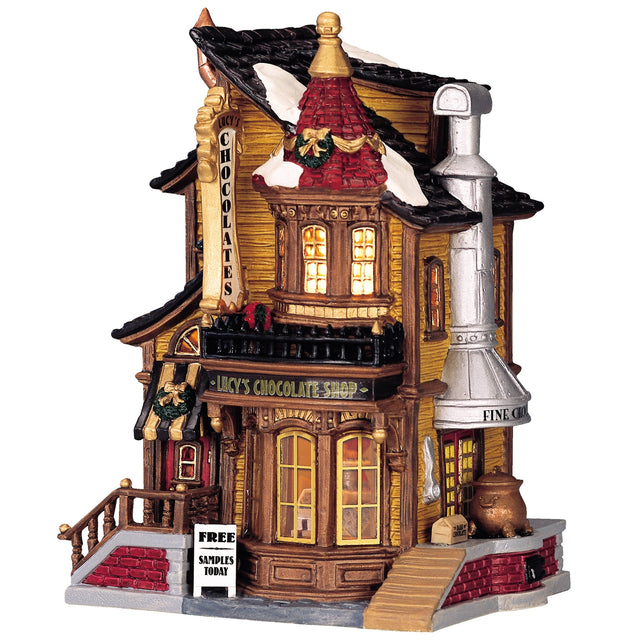 Porcelain lighted building for a Lemax display. Designed to look like an old Victorian-era house with wreaths and snow on it. The house is gold with brown frames. Inside the window you can see an array of chocolates.