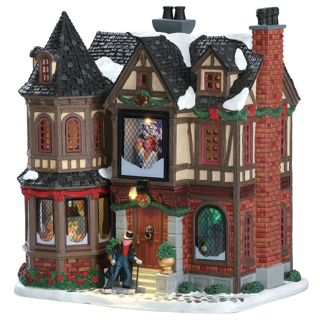 Lemax village building designed to look like an old Victorian style mansion covered in snow. There's green and red tinsel around the roof and some green bushes around the bottom. Scrooge is walking up the front steps.