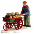 Polyresin figurine for a Lemax display. Designed to look like a food vendor pushing a red and green cart around in the snow. He's wearing a brown cowboy hat, a red scarf, a blue jacket, green pants and black shoes. The vendor's cart is full of pies and bread for sale.