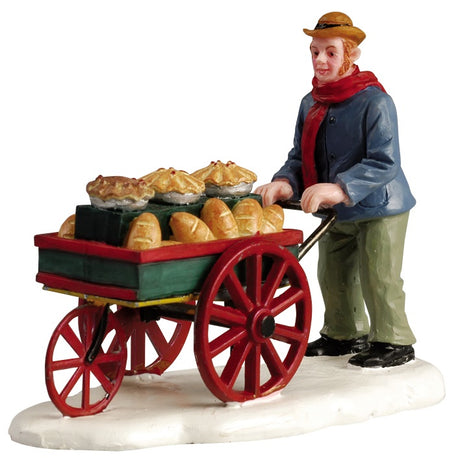 Polyresin figurine for a Lemax display. Designed to look like a food vendor pushing a red and green cart around in the snow. He's wearing a brown cowboy hat, a red scarf, a blue jacket, green pants and black shoes. The vendor's cart is full of pies and bread for sale.