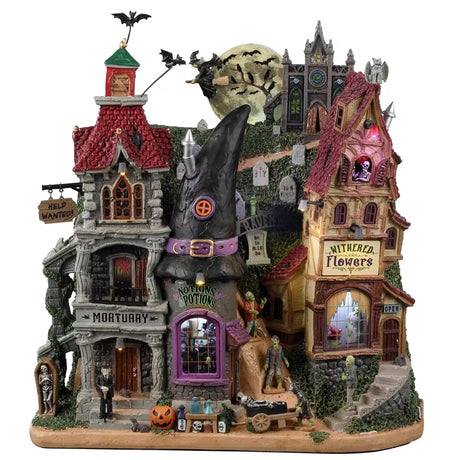 Resin building facade for a Spooky Town Lemax display. Designed to look like a bunch of store fronts. One section is a mortuary with a "help wanted" sign hanging from it. The building next to it reads "NOTION'S POTIONS" and has a witch hat for a roof. The building next to it is a flower shop. Behind them all is a cemetery and a spooky cathedral. There's a witch flying near the moon with bats following behind.