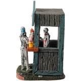 Resin village table accent for a Spooky Town Lemax display. Designed to look like a wooden candy stand. There's a skeleton serving candy to two young skeleton children. There are pots of candy on the side and pots of eyeballs on the other.