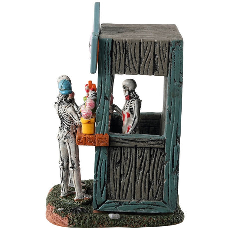 Resin village table accent for a Spooky Town Lemax display. Designed to look like a wooden candy stand. There's a skeleton serving candy to two young skeleton children. There are pots of candy on the side and pots of eyeballs on the other.