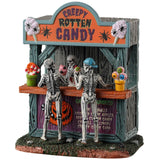 Resin village table accent for a Spooky Town Lemax display. Designed to look like a wooden candy stand. There's a skeleton serving candy to two young skeleton children. There are pots of candy on the side and pots of eyeballs on the other.