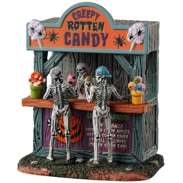 Resin village table accent for a Spooky Town Lemax display. Designed to look like a wooden candy stand. There's a skeleton serving candy to two young skeleton children. There are pots of candy on the side and pots of eyeballs on the other.