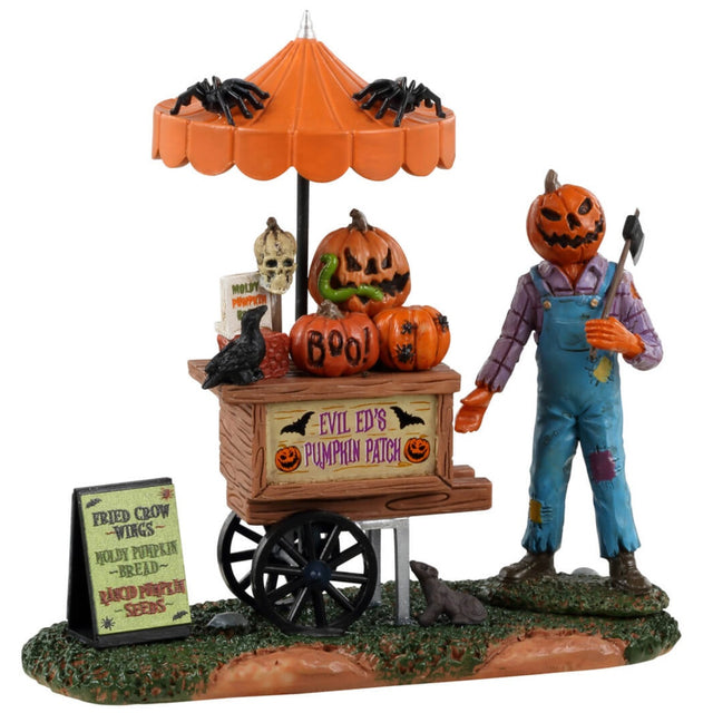 Resin table accent for a Lemax display. Designed to look like a scarecrow with a pumpkin head standing in front of a cart full of carved pumpkins. There's an orange umbrella above the cart with spiders on top. There's a sign on the ground that reads "Fried crow wings, moldy pumpkin bread and rancid pumpkin seeds."