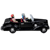 Resin village accent for a Lemax Spooky Town display. Designed to look like three people in a shiny, black convertible car. They're all dressed up. The driver is wearing a suit and a gray hat. The woman in the passenger seat is wearing a purple dress and a blue hat. The woman in the backseat is wearing a pink dress, a pearl necklace and a red hat. Their skin is green.