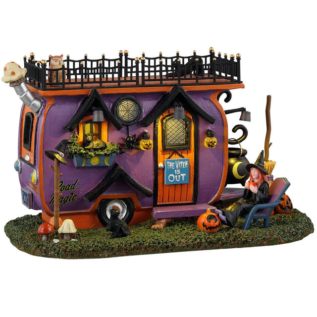 Resin village accent for a Lemax display. Designed to look like a purple, orange and black camper van parked on a patch of grass. There are mushrooms and carved pumpkins and black cats and an owl. There's a witch sitting in a blue chair.