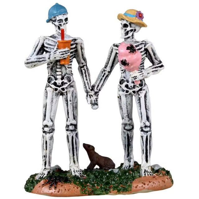 Resin figurine for a Lemax display. Designed to look like two skeletons holding hands. The one on the right is wearing a backwards blue baseball hat and holding an orange cup with a red straw. The one on the right is wearing a yellow hat with a flower on it and holding a stick of pink cotton candy with spiders on it. There's a large rat on the ground between them.